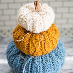 May include: A stack of three knitted pumpkins in blue, yellow, and white. The top pumpkin has a cinnamon stick stem.