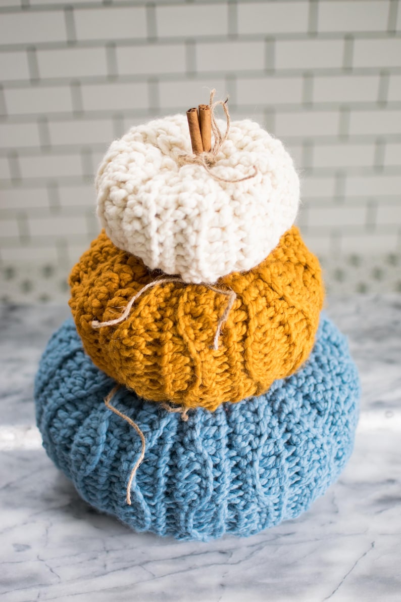 May include: A stack of three knitted pumpkins in blue, yellow, and white. The top pumpkin has a cinnamon stick stem.