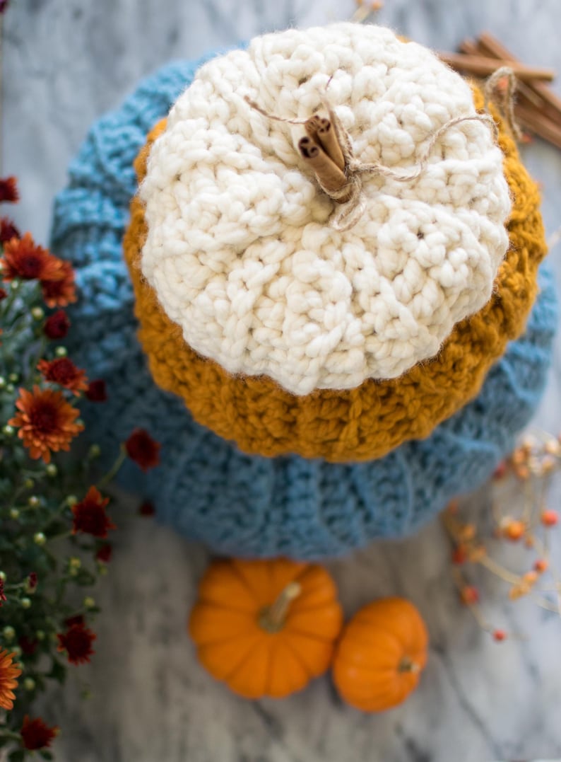 May include: A crocheted pumpkin decoration in blue, yellow, and white with cinnamon sticks and twine on top. Two small orange pumpkins are in the foreground.