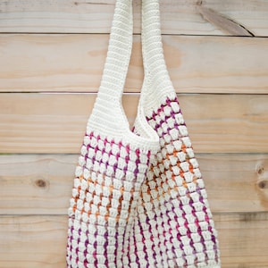 May include: A white crocheted tote bag with a pink, orange, and white striped pattern. The bag has a long strap and is hanging on a wooden wall.