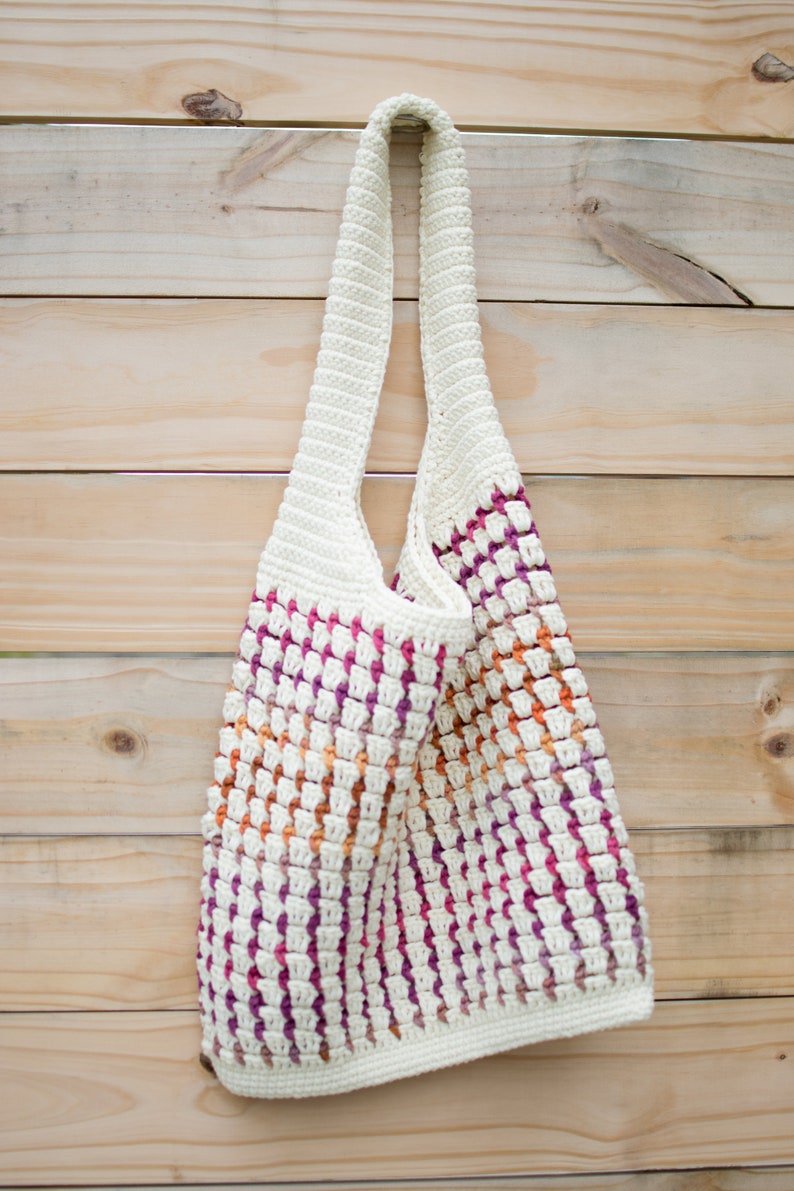 May include: A white crocheted tote bag with a pink, orange, and white striped pattern. The bag has a long strap and is hanging on a wooden wall.