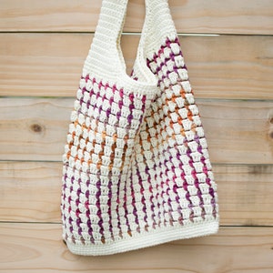 May include: A white crocheted tote bag with a colorful striped pattern in shades of pink, orange, and purple. The bag is hanging on a wooden surface.