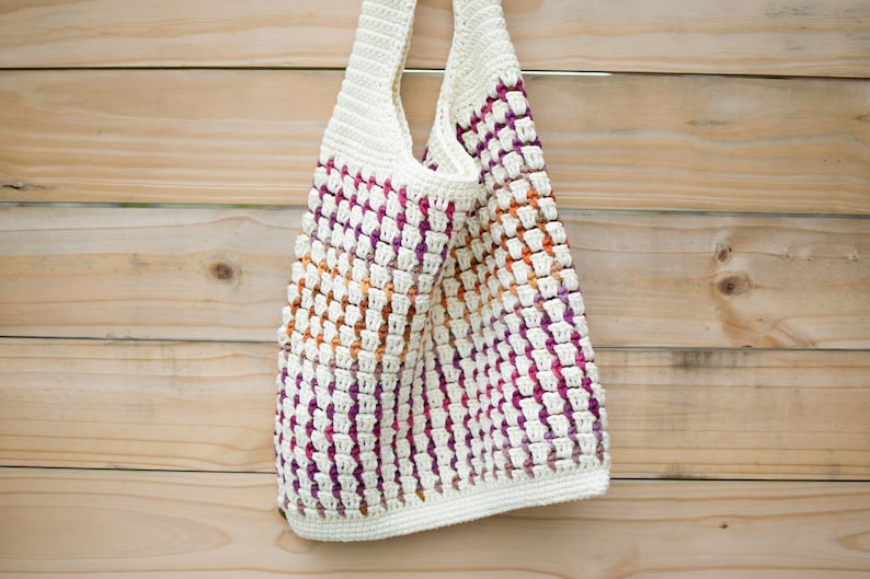 May include: A white crocheted tote bag with a colorful striped pattern in shades of pink, orange, and purple. The bag is hanging on a wooden surface.