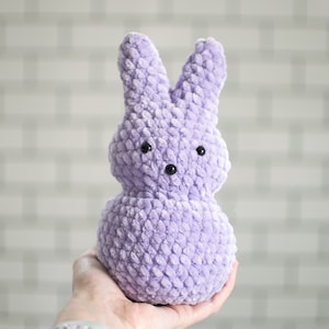 Puede incluir: Un conejito de peluche morado con ojos negros. El conejito está tejido a crochet y tiene una superficie texturizada.