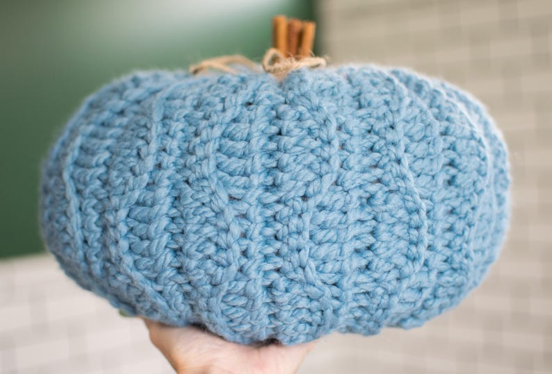 May include: A blue knitted pumpkin with a textured surface. The pumpkin is made of yarn and has a rustic, handmade look.