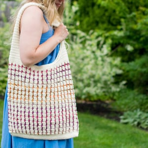 May include: A white crocheted tote bag with a colorful striped pattern. The bag is worn over the shoulder of a person wearing a blue dress.