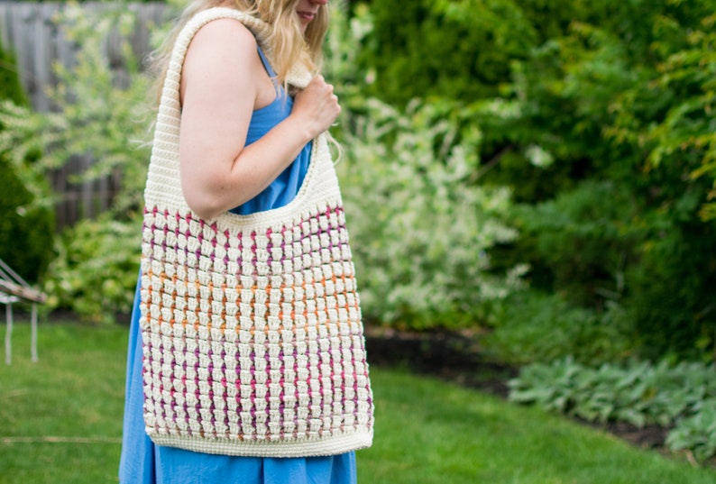 May include: A white crocheted tote bag with a colorful striped pattern. The bag is worn over the shoulder of a person wearing a blue dress.