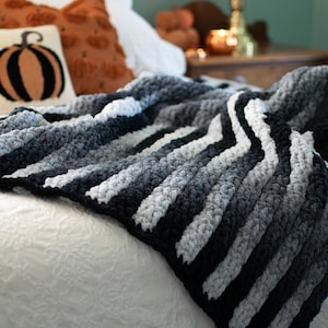 Könnte beinhalten: Eine kuschelige, gestreifte Decke in Grau-, Weiß- und Schwarztönen liegt auf einem Bett. Die grob gestrickte Decke hat ein strukturiertes Aussehen. Dekorative orangefarbene Kissen und ein Kürbiskissen sind im Hintergrund.
