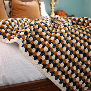 May include: A handmade crochet blanket in a granny square pattern. The blanket features a repeating pattern of white, navy blue, and mustard yellow squares. The blanket is draped over a bed with white linens and brown pillows.