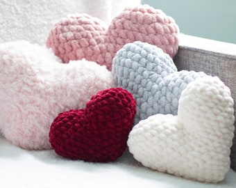 May include: Five heart-shaped pillows in various colors, including pink, white, red, and gray. The pillows are made of soft, fluffy material and are arranged on a white surface.