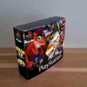 Puede incluir: Una caja rectangular de colección de videojuegos PlayStation con Crash Bandicoot en la portada. La ilustración muestra al personaje montando una motocicleta, con el logotipo de PlayStation y la etiqueta PAL visibles. El panel lateral enumera los títulos Crash Bandicoot 2 y Crash Bandicoot Warped.