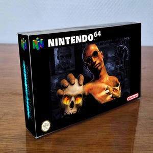 Peut inclure: Une boîte de jeu vidéo Nintendo 64 pour Shadow Man représentant un personnage chauve avec des lunettes rondes tenant un crâne aux yeux orange brillants. La boîte noire affiche le logo Nintendo 64 en haut et un sceau Nintendo dans le coin.