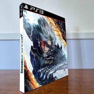 May include: A PlayStation 3 video game case for Metal Gear Rising: Revengeance. The cover art features a silver-haired character with a black eye patch and a metallic cybernetic arm, set against a background of orange flames and debris.