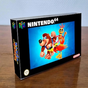 Op de afbeelding: Een Nintendo 64 videogame doos voor Banjo-Tooie met een zwarte rand, het Nintendo 64 logo linksboven en een centrale illustratie van een bruine beer, een rode vogel en een geel skeletachtig figuur tegen een helderblauwe achtergrond.