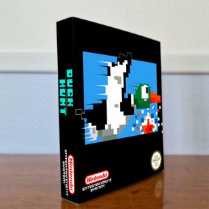 May include: A black rectangular video game box for Duck Hunt for the Nintendo Entertainment System. The front features a pixelated duck flying against a blue background. The side panel displays the title Duck Hunt in teal and white text.