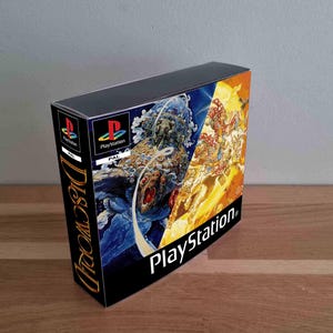 Puede incluir: Una caja rectangular de videojuego PlayStation para Discworld con un diseño de portada dividido en dos colores. El lado izquierdo muestra una escena cósmica azul oscuro con un elefante, mientras que el lado derecho presenta una ilustración de color amarillo y naranja brillante.