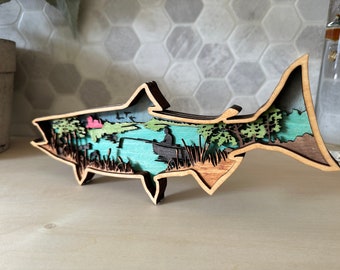 May include: A wooden cutout of a salmon with a detailed scene of a lake with a person fishing inside the fish's body. The scene is painted in shades of blue, green, and brown.