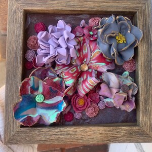 Könnte beinhalten: Ein gerahmtes Kunstwerk mit einer 3D-Blumenarrangements mit rosa, roten, violetten und blauen Blumen. Die Blumen sind aus Polymer Clay gefertigt und auf einem strukturierten Hintergrund angeordnet.