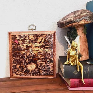 May include: A framed wood-burned art piece depicting a historical scene with figures and a castle. A gold fairy figurine with white wings sits on a stack of books next to a large mushroom sculpture. The art is displayed on a wooden surface.