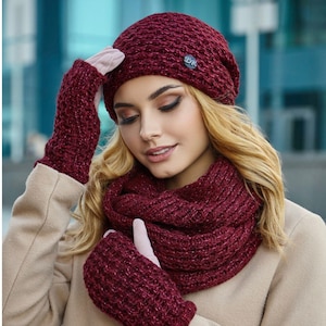Könnte beinhalten: Ein bordeauxfarbenes Strickmützen-, Schal- und fingerloses Handschuh-Set. Die Mütze hat einen kleinen dekorativen Knopf. Das Set ist aus einem strukturierten Strickmaterial gefertigt. Das Model trägt einen beigefarbenen Mantel.