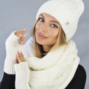 Może przedstawiać: Zestaw białego dzianinowego kapelusza, szalika i bezpalcowych rękawiczek. Czapka ma wzór warkocza i mały złoty guzik. Szalik i rękawiczki są również warkoczowe.