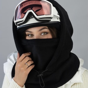 Könnte beinhalten: Ein schwarzer Fleece-Halswärmer mit Kordelzug. Der Halswärmer wird über einem weißen Skihelm mit einer rosafarbenen getönten Skibrille getragen.
