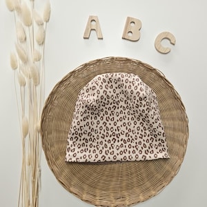 Könnte beinhalten: Eine beige Mütze mit braunem Leopardenmuster liegt auf einem runden Weidenkorb. Holzbuchstaben A, B und C befinden sich über dem Korb. Getrocknete weiße Blumen sind links.