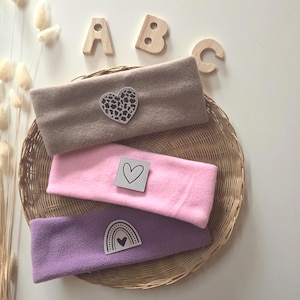 Könnte beinhalten: Drei weiche Stoffstirnbänder in Beige, Rosa und Lila. Das beige Stirnband hat einen herzförmigen Aufnäher mit Leopardenmuster. Das rosa Stirnband zeigt einen quadratischen Aufnäher mit Herzumriss. Das lila Stirnband ist mit einem Regenbogen-Aufnäher versehen.