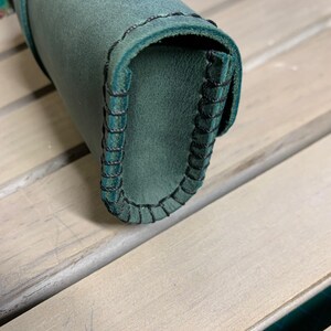 Puede incluir: Un estuche para l&aacute;pices de cuero verde oscuro con costuras negras. El estuche est&aacute; enrollado y las costuras son visibles en el borde.