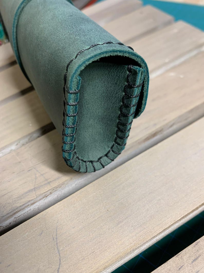Puede incluir: Un estuche para l&aacute;pices de cuero verde oscuro con costuras negras. El estuche est&aacute; enrollado y las costuras son visibles en el borde.