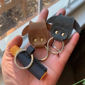 May include: Three leather keychains with a cow head design. The keychains are brown, black, and blue. The keychains are attached to silver key rings. The keychains are on a hand.
