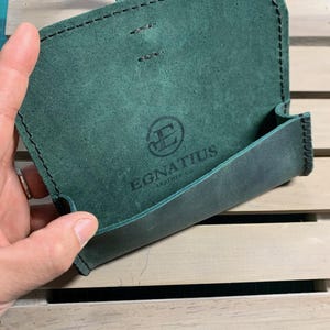 Puede incluir: Una funda de cuero verde con cierre de solapa y borde cosido. La funda tiene un logotipo que dice "Egnatius Leather Co."
