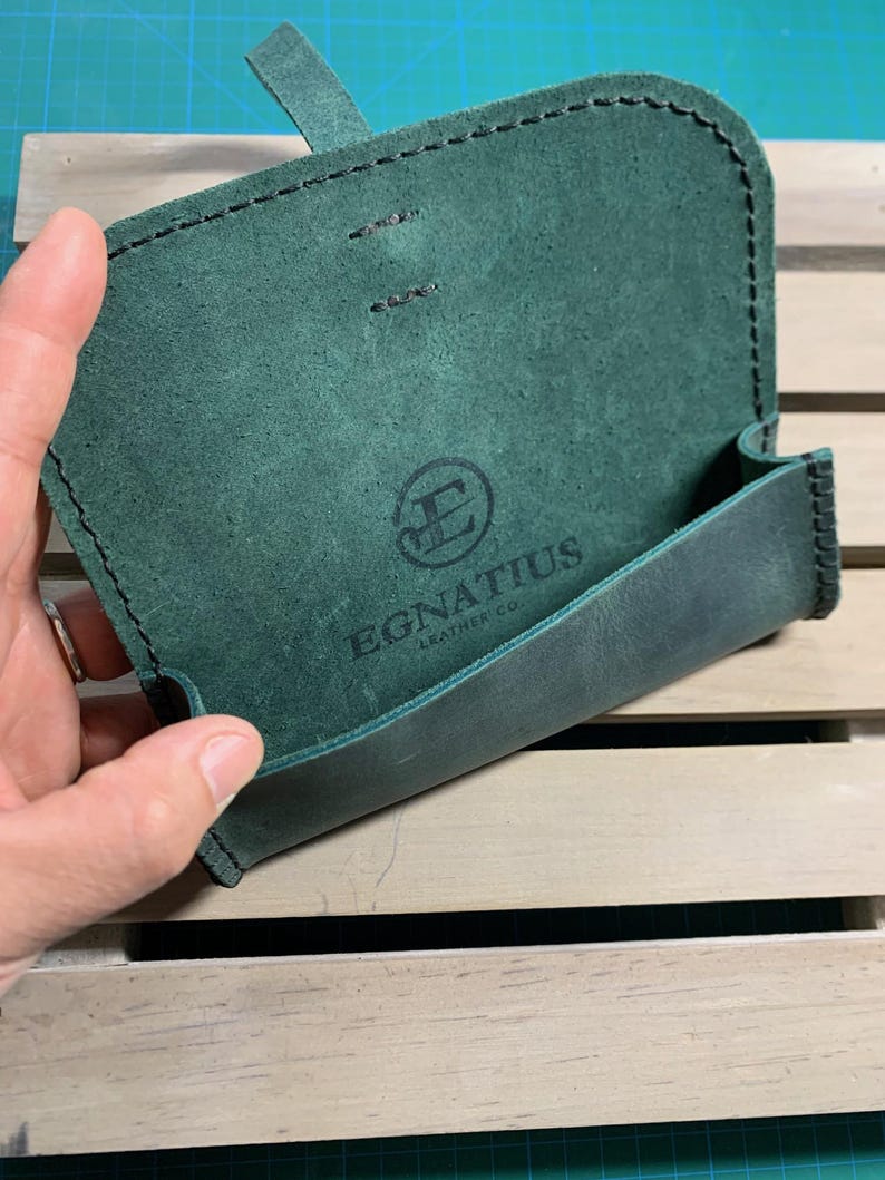 Puede incluir: Una funda de cuero verde con cierre de solapa y borde cosido. La funda tiene un logotipo que dice "Egnatius Leather Co."