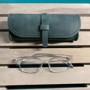 Puede incluir: Un estuche para gafas de cuero verde con cierre de correa. El estuche est&aacute; sobre una superficie de madera. Tambi&eacute;n se muestran gafas con montura transparente.