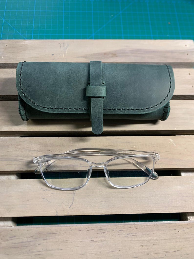 Puede incluir: Un estuche para gafas de cuero verde con cierre de correa. El estuche est&aacute; sobre una superficie de madera. Tambi&eacute;n se muestran gafas con montura transparente.