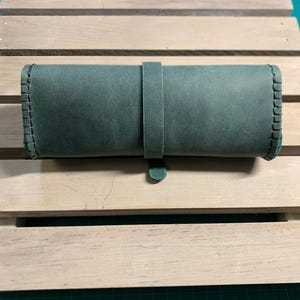 Puede incluir: Un estuche enrollable de cuero verde oscuro con un borde cosido y una correa de cuero.