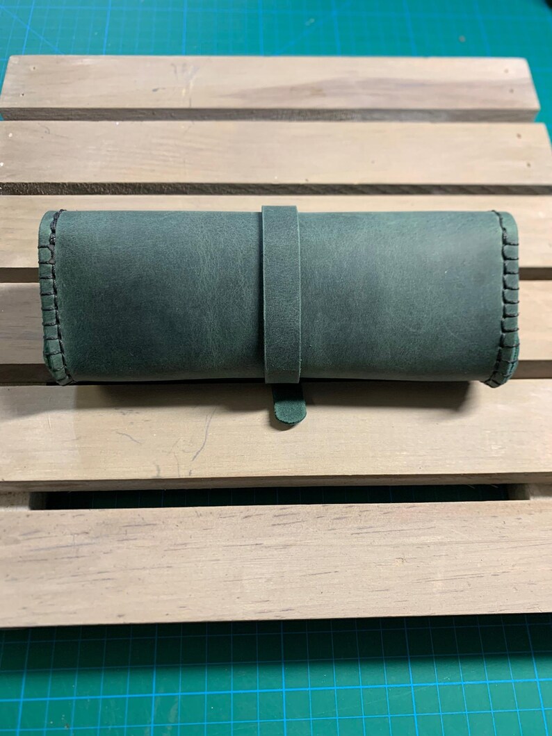 Puede incluir: Un estuche enrollable de cuero verde oscuro con un borde cosido y una correa de cuero.
