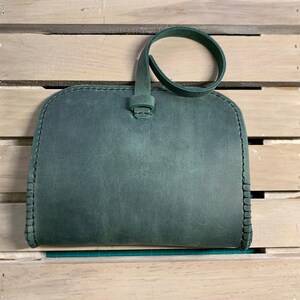 Puede incluir: Un clutch de cuero verde oscuro con una sola correa. El clutch tiene un borde cosido y una superficie lisa y texturizada.