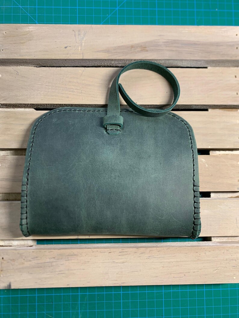 Puede incluir: Un clutch de cuero verde oscuro con una sola correa. El clutch tiene un borde cosido y una superficie lisa y texturizada.