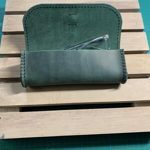 Puede incluir: Un estuche para gafas de cuero verde con un borde cosido y una solapa que se abre para revelar un par de gafas en el interior.
