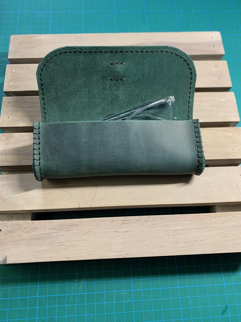 Puede incluir: Un estuche para gafas de cuero verde con un borde cosido y una solapa que se abre para revelar un par de gafas en el interior.