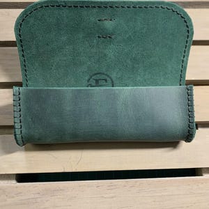 Puede incluir: Una funda de cuero verde con un borde cosido y un peque&ntilde;o logotipo en la parte delantera. La funda est&aacute; abierta y el interior est&aacute; forrado con un tejido verde suave.