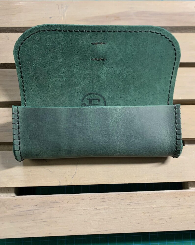 Puede incluir: Una funda de cuero verde con un borde cosido y un peque&ntilde;o logotipo en la parte delantera. La funda est&aacute; abierta y el interior est&aacute; forrado con un tejido verde suave.