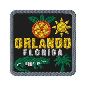 May include: A square embroidered patch with a black background and a grey border. It features a yellow orange slice with green leaves, a yellow sun, a green alligator, and a green palm tree. The text reads ORLANDO in yellow and FLORIDA in white.