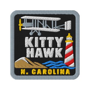 May include: A square embroidered patch with a grey border on a black background. It features a white biplane, a red and white striped lighthouse, tan sand dunes, and blue waves. The text reads KITTY HAWK and N. CAROLINA in white and yellow.