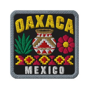 Può includere: Una toppa ricamata quadrata con sfondo nero e bordo grigio. Presenta il testo giallo OAXACA in alto e MEXICO in basso. Il centro mostra un vaso di terracotta marrone con motivi rossi e bianchi, una pianta di agave verde e un fiore rosso.