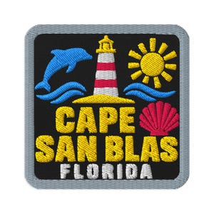 May include: A square embroidered patch with a black background and a gray border. It features a blue dolphin, a red and white lighthouse, a yellow sun, and a pink seashell. The text reads CAPE SAN BLAS in yellow and FLORIDA in white.