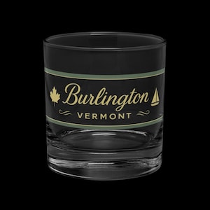 Puede incluir: Un vaso bajo transparente con una banda negra con el texto "Burlington Vermont" en letras doradas. El diseño incluye una hoja de arce dorada y un velero. El vaso está diseñado para beber.