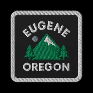 Puede incluir: Parche bordado con las palabras "EUGENE OREGON" en blanco, arqueadas sobre una montaña verde y dos pinos verdes. Un pequeño círculo gris está encima de la montaña. El parche tiene un fondo negro y un borde blanco.