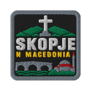 Puede incluir: Un parche bordado cuadrado con borde gris sobre fondo negro. Presenta una cruz blanca sobre montañas verdes, el texto blanco SKOPJE, el texto amarillo N MACEDONIA, un puente de piedra gris y un edificio gris con una franja roja y una aguja.